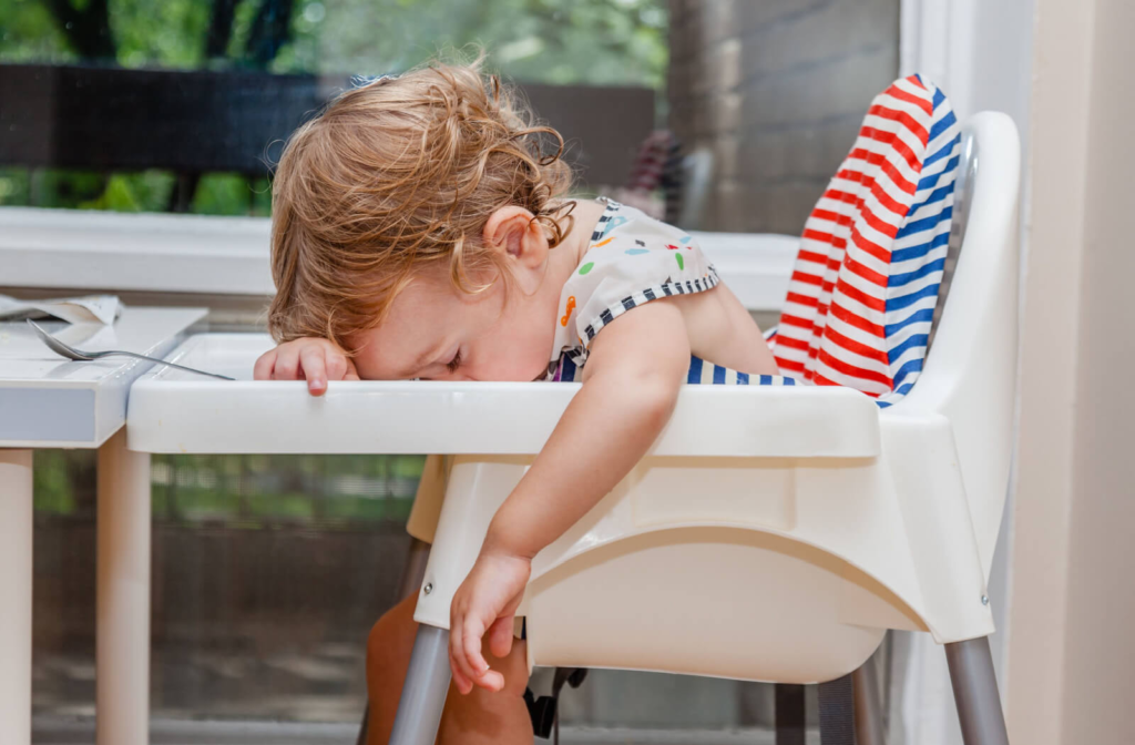 A sleeping baby in a high chair resting her head on the chair's attached table.