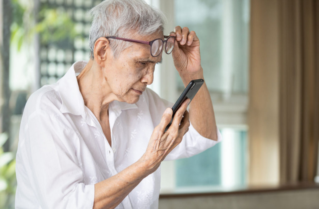 A senior woman squinting to see her cellphone as she holds her glasses above her eyes with her left hand.