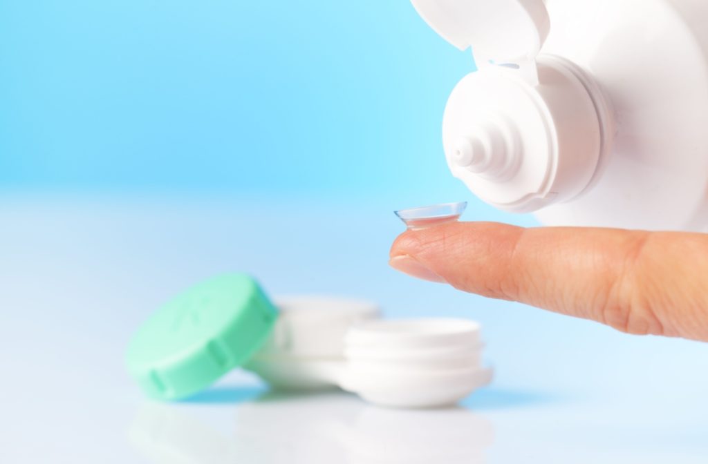 A single contact lens placed on a finger showing contact lens solution and a green and white contact lens case in the background.