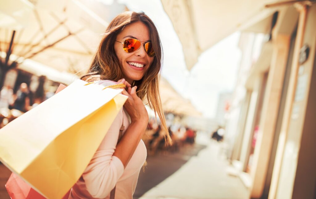 An adult stands in the sun and smiles while holding a shopping bag over their shoulder after a long day outside.